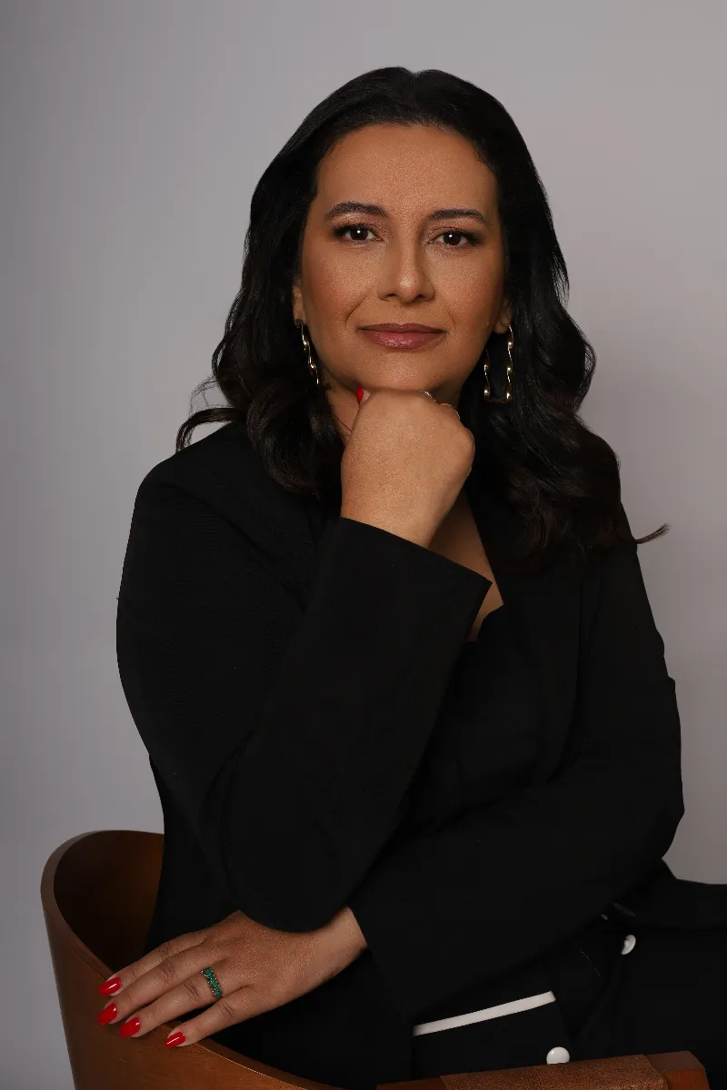 Retrato de Carol Villaça, de blazer preto, com a mão apoiada no queixo